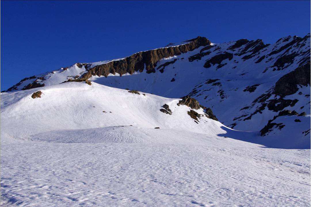 La selle neigeuse du Taou Bl : Pour rejoindre en NE le glacier de l'Aouille, remonter sous l'axe du plan incliné rocheux (casque recommandé / chutes de pierres). L'accès au versant N est le point lumineux à l'embase droite du bastion rocheux sommital